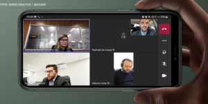 Montagem digital de um smartphone na mão de uma pessoa, na tela do equipamento é exibida uma reunião virtual da qual participa Alexandre Pires, o coordenador-geral do SINJUS e coordenador de Assuntos Jurídicos da Fenajud.