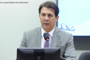 Deputado federal Arthur Maia, do Democratas da Bahia, sentado por detrás de uma mesa com um computador durante reunião na Câmara dos Deputados. Homem de pele clara, cabelo liso curto e castanho, trajando terno cinza, camisa branca e gravata roxa.