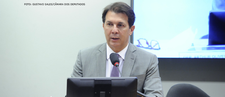 Deputado federal Arthur Maia, do Democratas da Bahia, sentado por detrás de uma mesa com um computador durante reunião na Câmara dos Deputados. Homem de pele clara, cabelo liso curto e castanho, trajando terno cinza, camisa branca e gravata roxa.