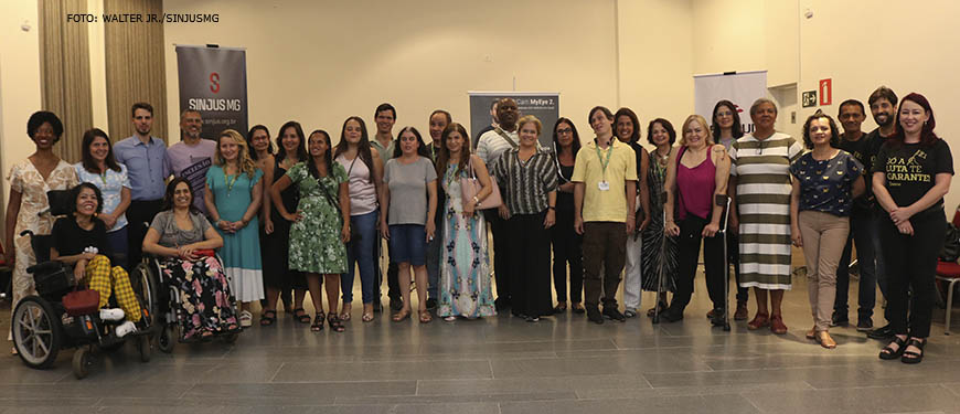 Fotografia com diversas pessoas, com e sem deficiência: são participantes e palestrantes do evento do núcleo da pessoa com deficiência do SINJUS e diretores do sindicato. O grupo está em um ambiente amplo, de paredes em tom creme e chão acinzentado; atrás das pessoas, há alguns banners.