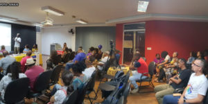 Um grupo de diversas pessoas, em sua maioria maioria negras, assistindo sentadas um homem negro falando ao microfone equanto mostra um slide com imagens sobre um telão.