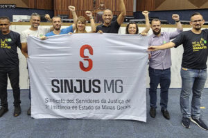 Imagem Acessível: Diretoria colegiada do SINJUS posa junto ao vereador e diretor de Assuntos Jurídicos Wagner Ferreira. Eles estão com a bandeira do Sindicato no plenário da Câmara Municipal de Belo Horizonte, todos sorriem e estão com braços para o alto com os punhos cerrados em um gesto de luta.