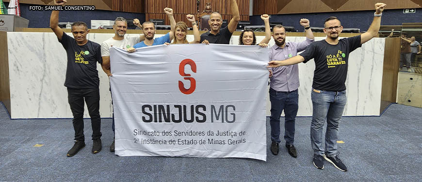 Imagem Acessível: Diretoria colegiada do SINJUS posa junto ao vereador e diretor de Assuntos Jurídicos Wagner Ferreira. Eles estão com a bandeira do Sindicato no plenário da Câmara Municipal de Belo Horizonte, todos sorriem e estão com braços para o alto com os punhos cerrados em um gesto de luta.