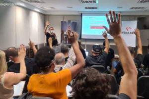 Foto de pessoas sentadas em um auditório com as mãos para cima em sinal de aprovação. A frente o Coordenador Geral do Sinjus-MG Alexandre Pires (homem de pele clara, cabelos curtos e castanhos usando uma blusa na cor preta, segurando um microfone fazendo o mesmo gesto da plateia.)