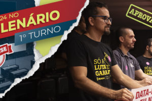 A imagem mostra diretores do SINJUS-MG acompanhando a votação na ALMG, vistos em um mezanino do plenário. Eles vestem camisetas do SINJUS-MG com a frase “Só a luta te garante” e seguram cartazes com mensagens como “Data-Base eu não abro mão!” e “Data-Base já!”. Todos estão concentrados, olhando em direção ao plenário.