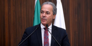 Foto de um homem falando ao microfone em um ambiente institucional. Ele aparenta ser de meia-idade, com pele clara, cabelos curtos e grisalhos, e expressão séria e concentrada, olhando levemente para o lado. Está vestido com terno escuro, camisa branca e gravata vermelha com padrão detalhado. À sua frente, há dois microfones posicionados em um púlpito, indicando que ele está fazendo um discurso. Ao fundo, há uma parede de madeira com acabamento vertical, além de duas bandeiras parcialmente visíveis, a do Brasil e a de Minas Gerais, reforçando o contexto formal e jurídico.