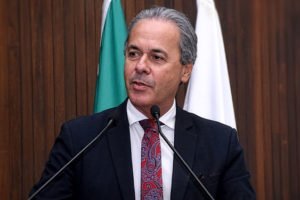 Foto de um homem falando ao microfone em um ambiente institucional. Ele aparenta ser de meia-idade, com pele clara, cabelos curtos e grisalhos, e expressão séria e concentrada, olhando levemente para o lado. Está vestido com terno escuro, camisa branca e gravata vermelha com padrão detalhado. À sua frente, há dois microfones posicionados em um púlpito, indicando que ele está fazendo um discurso. Ao fundo, há uma parede de madeira com acabamento vertical, além de duas bandeiras parcialmente visíveis, a do Brasil e a de Minas Gerais, reforçando o contexto formal e jurídico.