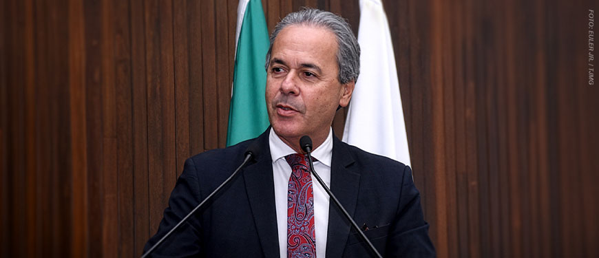 Foto de um homem falando ao microfone em um ambiente institucional. Ele aparenta ser de meia-idade, com pele clara, cabelos curtos e grisalhos, e expressão séria e concentrada, olhando levemente para o lado. Está vestido com terno escuro, camisa branca e gravata vermelha com padrão detalhado. À sua frente, há dois microfones posicionados em um púlpito, indicando que ele está fazendo um discurso. Ao fundo, há uma parede de madeira com acabamento vertical, além de duas bandeiras parcialmente visíveis, a do Brasil e a de Minas Gerais, reforçando o contexto formal e jurídico.