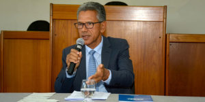 Homem sentado em uma mesa, falando ao microfone em ambiente institucional. Ele aparenta ser de meia-idade, tem pele morena, cabelos curtos grisalhos e usa óculos de armação escura. Sua expressão é séria e engajada, com o olhar direcionado à frente. Ele está vestido com terno escuro, camisa clara e gravata azul com pequenos padrões. Com uma das mãos, segura o microfone próximo à boca; com a outra, faz um gesto aberto, como se estivesse enfatizando um ponto durante a fala.