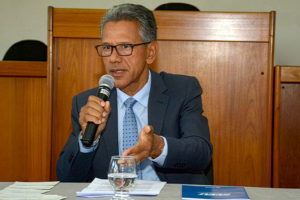 Homem sentado em uma mesa, falando ao microfone em ambiente institucional. Ele aparenta ser de meia-idade, tem pele morena, cabelos curtos grisalhos e usa óculos de armação escura. Sua expressão é séria e engajada, com o olhar direcionado à frente. Ele está vestido com terno escuro, camisa clara e gravata azul com pequenos padrões. Com uma das mãos, segura o microfone próximo à boca; com a outra, faz um gesto aberto, como se estivesse enfatizando um ponto durante a fala.