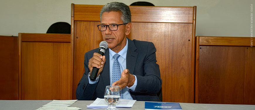 Homem sentado em uma mesa, falando ao microfone em ambiente institucional. Ele aparenta ser de meia-idade, tem pele morena, cabelos curtos grisalhos e usa óculos de armação escura. Sua expressão é séria e engajada, com o olhar direcionado à frente. Ele está vestido com terno escuro, camisa clara e gravata azul com pequenos padrões. Com uma das mãos, segura o microfone próximo à boca; com a outra, faz um gesto aberto, como se estivesse enfatizando um ponto durante a fala.