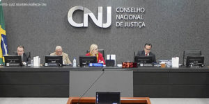 Mesa diretiva do CNJ durante sessão ordinária com a presidente do órgão, ministra Rosa Weber. Ao fundo está um grande letreiro em aço inox com fundo acarpetado cinza escuro, nele está escrito CNJ - Conselho Nacional de Justiça.