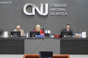 Mesa diretiva do CNJ durante sessão ordinária com a presidente do órgão, ministra Rosa Weber. Ao fundo está um grande letreiro em aço inox com fundo acarpetado cinza escuro, nele está escrito CNJ - Conselho Nacional de Justiça.