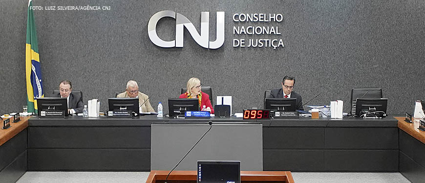 Mesa diretiva do CNJ durante sessão ordinária com a presidente do órgão, ministra Rosa Weber. Ao fundo está um grande letreiro em aço inox com fundo acarpetado cinza escuro, nele está escrito CNJ - Conselho Nacional de Justiça.