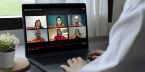 Pessoa usando um notebook, assistindo a uma videoconferência com seis participantes, organizada em dois blocos de três telas. A reunião virtual aborda o tema “ASSÉDIO MORAL - Prevenção e Combate”. No canto superior esquerdo, aparece Luciane Santos, Feserv Minas, ao lado de Fanny Melo, SINDSEMP-MG / Feserv Minas” e Eduardo Maia, SINDSEMP-MG. No bloco inferior, à esquerda, está Wagner Ferreira, SINJUS-MG/ Feserv Minas, ao centro Mariana Tavares, Advogada, e à direita Aline, interprete de Libras. A pessoa que assiste está sentada à mesa, com a mão apoiada no teclado. Ao fundo, uma janela com cortinas brancas.