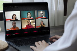 Pessoa usando um notebook, assistindo a uma videoconferência com seis participantes, organizada em dois blocos de três telas. A reunião virtual aborda o tema “ASSÉDIO MORAL - Prevenção e Combate”. No canto superior esquerdo, aparece Luciane Santos, Feserv Minas, ao lado de Fanny Melo, SINDSEMP-MG / Feserv Minas” e Eduardo Maia, SINDSEMP-MG. No bloco inferior, à esquerda, está Wagner Ferreira, SINJUS-MG/ Feserv Minas, ao centro Mariana Tavares, Advogada, e à direita Aline, interprete de Libras. A pessoa que assiste está sentada à mesa, com a mão apoiada no teclado. Ao fundo, uma janela com cortinas brancas.