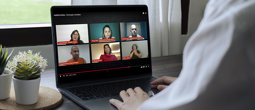 Pessoa usando um notebook, assistindo a uma videoconferência com seis participantes, organizada em dois blocos de três telas. A reunião virtual aborda o tema “ASSÉDIO MORAL - Prevenção e Combate”. No canto superior esquerdo, aparece Luciane Santos, Feserv Minas, ao lado de Fanny Melo, SINDSEMP-MG / Feserv Minas” e Eduardo Maia, SINDSEMP-MG. No bloco inferior, à esquerda, está Wagner Ferreira, SINJUS-MG/ Feserv Minas, ao centro Mariana Tavares, Advogada, e à direita Aline, interprete de Libras. A pessoa que assiste está sentada à mesa, com a mão apoiada no teclado. Ao fundo, uma janela com cortinas brancas.