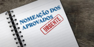 imagem mostra um caderno de espiral aberto, com as palavras “Nomeação dos Aprovados” escritas em letras grandes e azuis, acompanhadas de um carimbo vermelho com a palavra “Urgente”.
