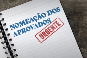 imagem mostra um caderno de espiral aberto, com as palavras “Nomeação dos Aprovados” escritas em letras grandes e azuis, acompanhadas de um carimbo vermelho com a palavra “Urgente”.