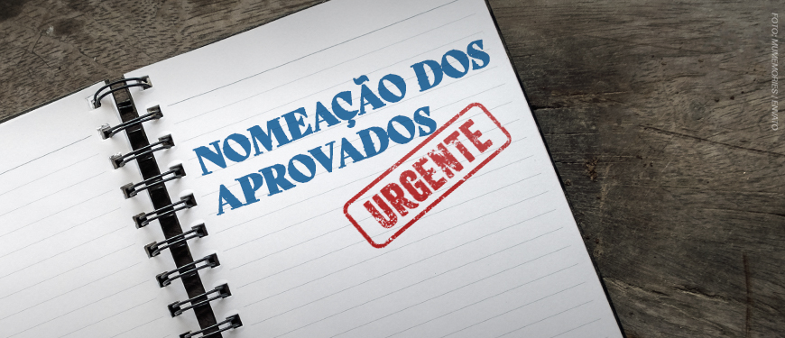 imagem mostra um caderno de espiral aberto, com as palavras “Nomeação dos Aprovados” escritas em letras grandes e azuis, acompanhadas de um carimbo vermelho com a palavra “Urgente”.