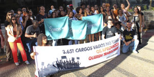 A imagem mostra um grupo numeroso de pessoas reunidas em frente à sede do TJMG, participando de um ato público. Os manifestantes, identificados como aprovados no concurso do Edital 1/2022, seguram faixas e cartazes que denunciam a existência de 3.651 cargos vagos e exigem nomeações. Muitos dos participantes estão com camisetas de entidades de classe, como o SERJUSMIG e o CRESS-MG, e demonstram engajamento e mobilização. O texto sobreposto à imagem informa: “Comissão de aprovados do concurso do Edital 1/2022 promove ato cobrando nomeações".
