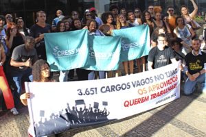 A imagem mostra um grupo numeroso de pessoas reunidas em frente à sede do TJMG, participando de um ato público. Os manifestantes, identificados como aprovados no concurso do Edital 1/2022, seguram faixas e cartazes que denunciam a existência de 3.651 cargos vagos e exigem nomeações. Muitos dos participantes estão com camisetas de entidades de classe, como o SERJUSMIG e o CRESS-MG, e demonstram engajamento e mobilização. O texto sobreposto à imagem informa: “Comissão de aprovados do concurso do Edital 1/2022 promove ato cobrando nomeações".