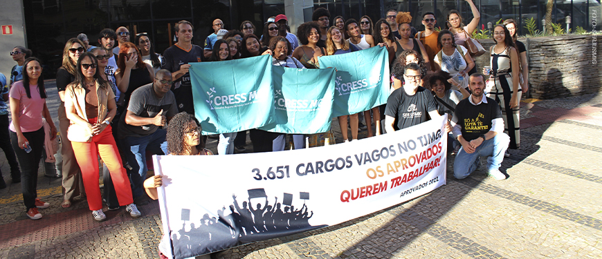 A imagem mostra um grupo numeroso de pessoas reunidas em frente à sede do TJMG, participando de um ato público. Os manifestantes, identificados como aprovados no concurso do Edital 1/2022, seguram faixas e cartazes que denunciam a existência de 3.651 cargos vagos e exigem nomeações. Muitos dos participantes estão com camisetas de entidades de classe, como o SERJUSMIG e o CRESS-MG, e demonstram engajamento e mobilização. O texto sobreposto à imagem informa: “Comissão de aprovados do concurso do Edital 1/2022 promove ato cobrando nomeações".