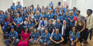 A imagem retrata um grande grupo de servidores que participaram do seminário, reunidos em uma sala, muitos deles vestindo camisetas azuis com a logo da identidade do evento, em pose para a foto em uma demonstração de diversidade e inclusão. O texto em destaque ressalta que o encontro reuniu pessoas com deficiência que enfrentam barreiras físicas e atitudinais no TJMG e no MPMG.