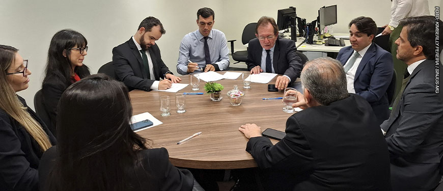 Imagem Acessível: Reunião em uma sala com pessoas sentadas ao redor de uma mesa redonda, estão presentes representantes do Serjusmig, Sindojus/MG, o presidente do TJMMG, Jadir Silva, o procurador do Estado, Daniel Cabalero Saldanha, o desembargador do TJMG, Paulo Mendes Álvares e o diretor de assuntos Jurídicos do SINJUS-MG, Felipe Rodrigues, que está assinando um documento. Conteúdo textual: Conquista - Servidores do TJM vão receber juros e correção das Datas-Bases de 2014 a 2017.