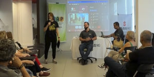 Imagem acessível: Fotografia do encontro promovido pelo Núcleo de Aposentados e Pensionistas (NAP) do SINJUS-MG, para debater direitos e reivindicações dos aposentados. Em uma sala com participantes sentados em semicírculo, a diretora Janaína Torres fala ao microfone, enquanto o coordenador-geral Felipe Rodrigues acompanha a reunião sentado ao centro. Ao fundo, uma tela exibe a transmissão online do encontro.