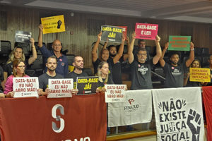 A imagem mostra um grupo de servidoras e servidores mobilizados em plenário, segurando cartazes com frases como “DATA-BASE JÁ!”, “DATA-BASE É LEI!” e “DATA-BASE EU NÃO ABRO MÃO!”. À frente, está estendida uma faixa do SINJUS-MG – Sindicato dos Servidores da Justiça de 2ª Instância do Estado de Minas Gerais, além de bandeiras de entidades sindicais. No topo da imagem, aparece a editoria “MOMENTO DECISIVO” e, em destaque, o texto: “Data-Base 2025 é aprovada em 1º turno e já volta à pauta às 10h desta 4ªf.”