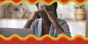 Imagem Acessível: Mulher negra com tranças longas está em frente a um computador, ela está levantando os óculos e massageia a testa, sinalizando cansaço