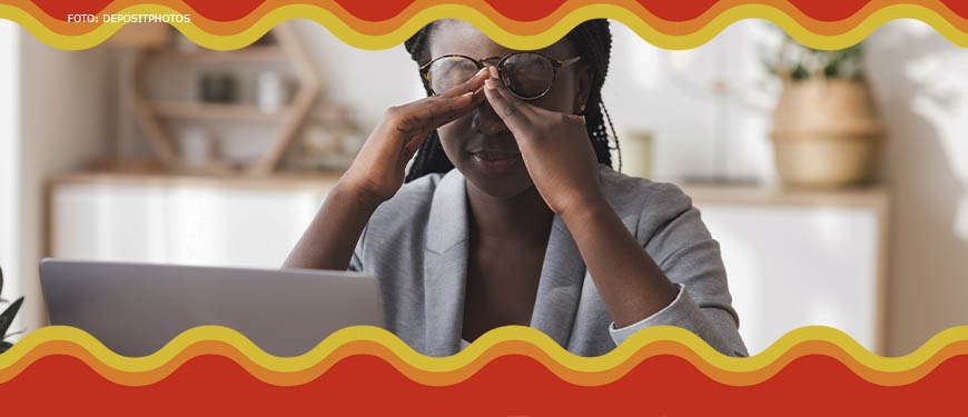 Imagem Acessível: Mulher negra com tranças longas está em frente a um computador, ela está levantando os óculos e massageia a testa, sinalizando cansaço