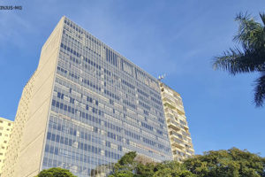 Fotografia diurna com vista frontal da unidade do TJMG, um prédio na cor branca com muitas janelas, onde se vê a sua frente uma rua movimentada com carros e arborizada.