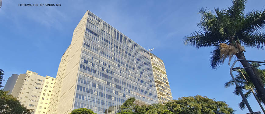 Fotografia diurna com vista frontal da unidade do TJMG, um prédio na cor branca com muitas janelas, onde se vê a sua frente uma rua movimentada com carros e arborizada.