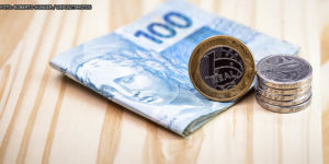em um fundo de madeira clara está um pequeno maço de dinheiro envolto por uma nota de cem reais, por cima deste conjunto está uma pilha de moedas de um real e de cinquenta centavos.