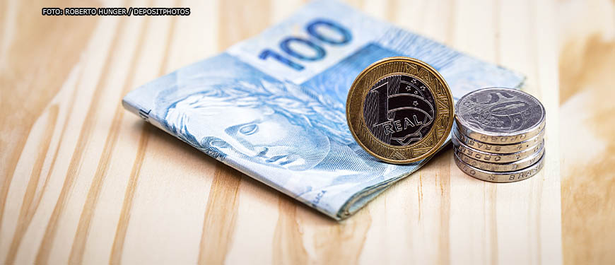 em um fundo de madeira clara está um pequeno maço de dinheiro envolto por uma nota de cem reais, por cima deste conjunto está uma pilha de moedas de um real e de cinquenta centavos.