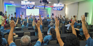 imagem mostra um auditório cheio, onde servidores com deficiência participam do seminário intersindical, todos vestidos com camisetas azuis e com as mãos levantadas em votação. No palco, representantes conduzem a atividade em frente a uma tela projetada, com bandeiras ao lado.