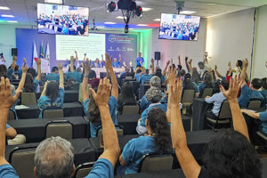 imagem mostra um auditório cheio, onde servidores com deficiência participam do seminário intersindical, todos vestidos com camisetas azuis e com as mãos levantadas em votação. No palco, representantes conduzem a atividade em frente a uma tela projetada, com bandeiras ao lado.