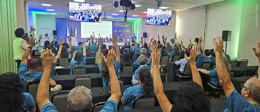 imagem mostra um auditório cheio, onde servidores com deficiência participam do seminário intersindical, todos vestidos com camisetas azuis e com as mãos levantadas em votação. No palco, representantes conduzem a atividade em frente a uma tela projetada, com bandeiras ao lado.