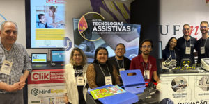 Foto de grupo nos estandes da Mostra de Tecnologias Assistivas e Soluções de Acessibilidade, promovida pelo SINJUS-MG. À esquerda está José Carlos Tonolli, da Click Tecnologia Assistiva, ao lado de equipamentos, notebooks e materiais de comunicação da empresa. No centro, a equipe da UFU é formada por Eryc Dias, Denize Novais, Dayane Nascimento e Marco Túlio Rodrigues, posicionados diante de uma tela com a identidade visual da Mostra e junto a dispositivos e recursos de tecnologia assistiva. À direita, a equipe da UFMG reúne Sarah Cortezzi, Jorge Bençussan e Pedro Caversan, atrás de uma bancada com protótipos, equipamentos eletrônicos e materiais relacionados a tecnologia assistiva e biomecânica. Todos posam para a foto no espaço da exposição.