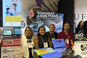 Foto de grupo nos estandes da Mostra de Tecnologias Assistivas e Soluções de Acessibilidade, promovida pelo SINJUS-MG. À esquerda está José Carlos Tonolli, da Click Tecnologia Assistiva, ao lado de equipamentos, notebooks e materiais de comunicação da empresa. No centro, a equipe da UFU é formada por Eryc Dias, Denize Novais, Dayane Nascimento e Marco Túlio Rodrigues, posicionados diante de uma tela com a identidade visual da Mostra e junto a dispositivos e recursos de tecnologia assistiva. À direita, a equipe da UFMG reúne Sarah Cortezzi, Jorge Bençussan e Pedro Caversan, atrás de uma bancada com protótipos, equipamentos eletrônicos e materiais relacionados a tecnologia assistiva e biomecânica. Todos posam para a foto no espaço da exposição.