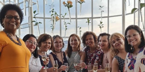 Fotografia da confraternização do Núcleo das Mulheres com o semblante de contentamento, no terraço do Edifício Acaiaca, ao fundo se vê a janela com a paisagem do céu.