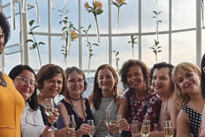 Fotografia da confraternização do Núcleo das Mulheres com o semblante de contentamento, no terraço do Edifício Acaiaca, ao fundo se vê a janela com a paisagem do céu.