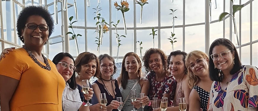 Fotografia da confraternização do Núcleo das Mulheres com o semblante de contentamento, no terraço do Edifício Acaiaca, ao fundo se vê a janela com a paisagem do céu.