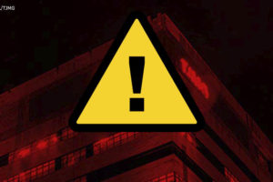 Foto da Sede do Tribunal de Justiça de Minas Gerais com um filtro vermelho-escuro aplicado sobre ela; sobre o edifício há um símbolo triangular de alerta, com fundo amarelo e um sinal de exclamação preto no centro.