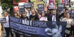 Imagem Acessível: A imagem mostra um grupo de pessoas reunidas em ato público em defesa das crianças. Elas seguram faixas e cartazes com mensagens como “Criança não é esposa”, “Infância é para brincar, não para casar” e “Exploração sexual infantil é crime hediondo”. Na frente, uma grande faixa do Núcleo das Mulheres (NM) do SINJUS-MG reforça o combate à exploração sexual infantil. O texto da arte destaca: “NM se mobiliza em ato pela proteção das crianças”, evidenciando a participação do núcleo na mobilização contra a pedofilia e a violência sexual.