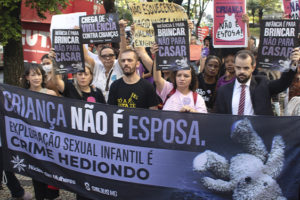 Imagem Acessível: A imagem mostra um grupo de pessoas reunidas em ato público em defesa das crianças. Elas seguram faixas e cartazes com mensagens como “Criança não é esposa”, “Infância é para brincar, não para casar” e “Exploração sexual infantil é crime hediondo”. Na frente, uma grande faixa do Núcleo das Mulheres (NM) do SINJUS-MG reforça o combate à exploração sexual infantil. O texto da arte destaca: “NM se mobiliza em ato pela proteção das crianças”, evidenciando a participação do núcleo na mobilização contra a pedofilia e a violência sexual.