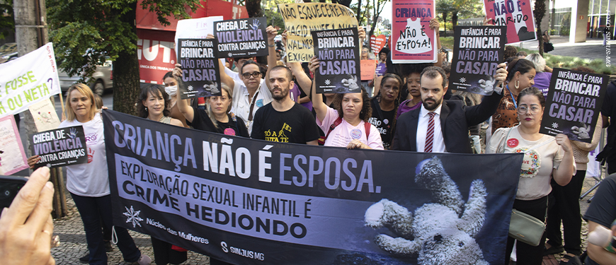Imagem Acessível: A imagem mostra um grupo de pessoas reunidas em ato público em defesa das crianças. Elas seguram faixas e cartazes com mensagens como “Criança não é esposa”, “Infância é para brincar, não para casar” e “Exploração sexual infantil é crime hediondo”. Na frente, uma grande faixa do Núcleo das Mulheres (NM) do SINJUS-MG reforça o combate à exploração sexual infantil. O texto da arte destaca: “NM se mobiliza em ato pela proteção das crianças”, evidenciando a participação do núcleo na mobilização contra a pedofilia e a violência sexual.