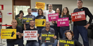 A imagem mostra um grupo de servidoras e servidores mobilizados em ambiente interno, segurando cartazes com mensagens como “DATA-BASE JÁ!”, “DATA-BASE É LEI!”, “DATA-BASE EU NÃO ABRO MÃO!” e “DATA-BASE REPONDO A INFLAÇÃO!”. No topo, aparece a editoria “AVANÇO” e, em destaque, o texto: “Data-Base 2025 é aprovada na FFO; votação no Plenário pode acontecer nesta quinta-feira.” A cena reforça o clima de mobilização e pressão pela recomposição inflacionária e pela aprovação definitiva da data-base.